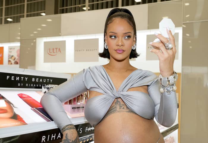 Rihanna holding a compact and standing next to a Fenty Beauty shelf in a makeup store