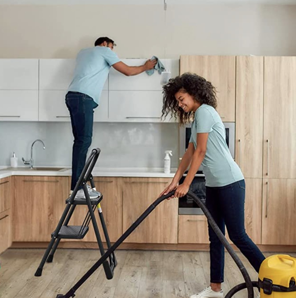 A man on the 2-step ladder in the kitchen with a woman cleaning floors