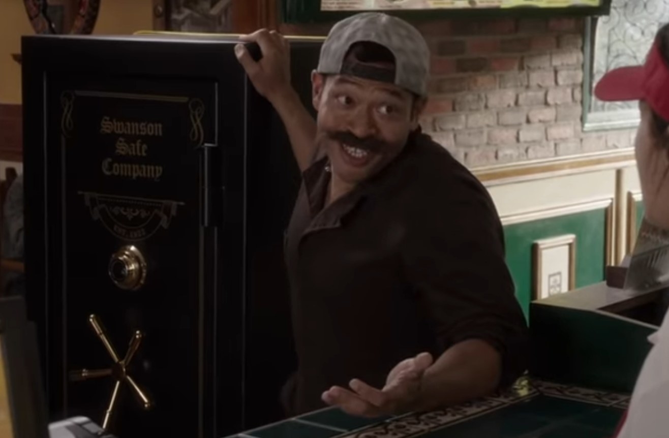 Eugene Cordero as Pillboi, wearing a fake mustache and a backwards baseball cap, standing in front of a safe