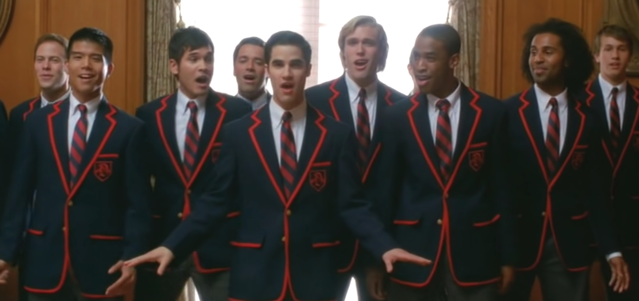 Blaine Anderson stands in front of the Warblers boys choir as they all wear matching dark colored blazers