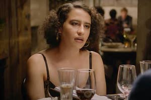 Ilana from Broad City staring blankly ahead at a fancy restaurant table with wine glasses in front of her