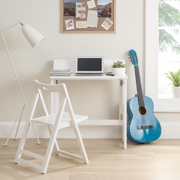 White wooden desk with laptop, books and planter, matching white chair, white lamp on left, blue guitar on right