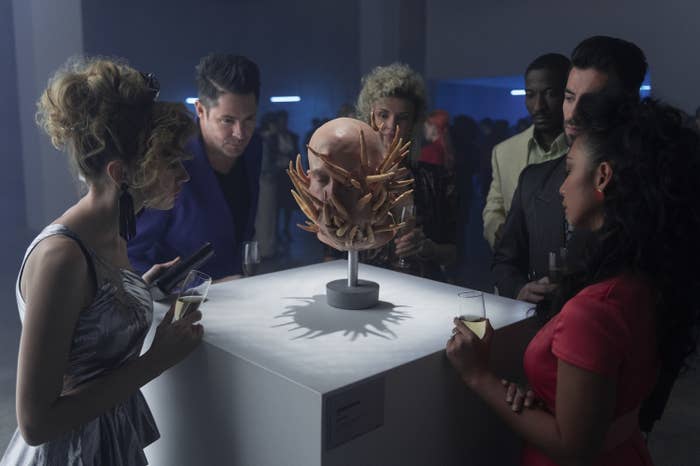 A group of people gather around an art piece composed of a human head with fingers jutting out from it