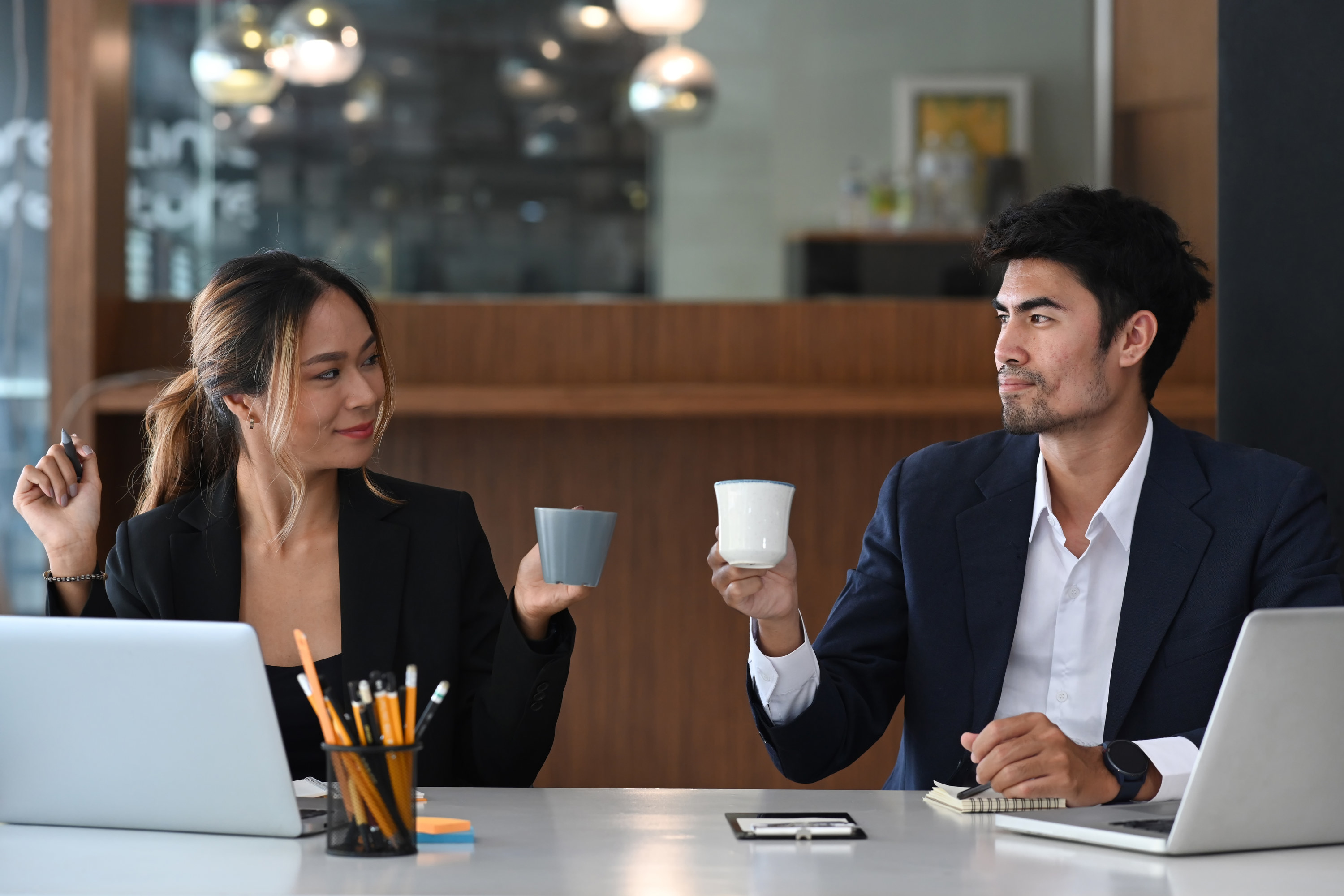 Man and woman sitting at a desk holding up their coffee mugs