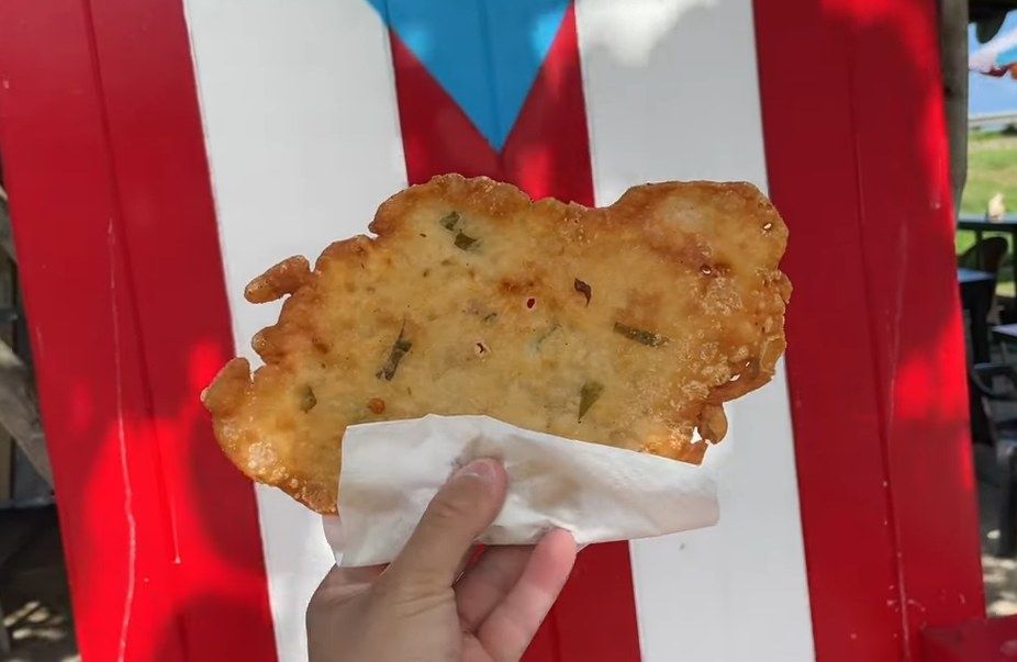 A hand holding a bacalaito in a napkin in front of a lounge chair with a Puerto Rican flag on it