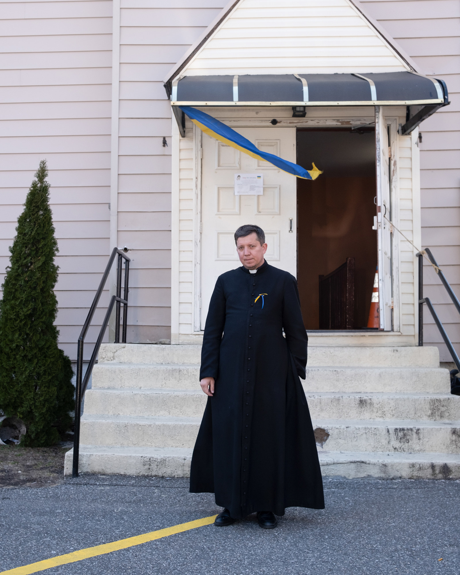 A priest stands in front of his church; a Ukrainian flag waves in front of the door