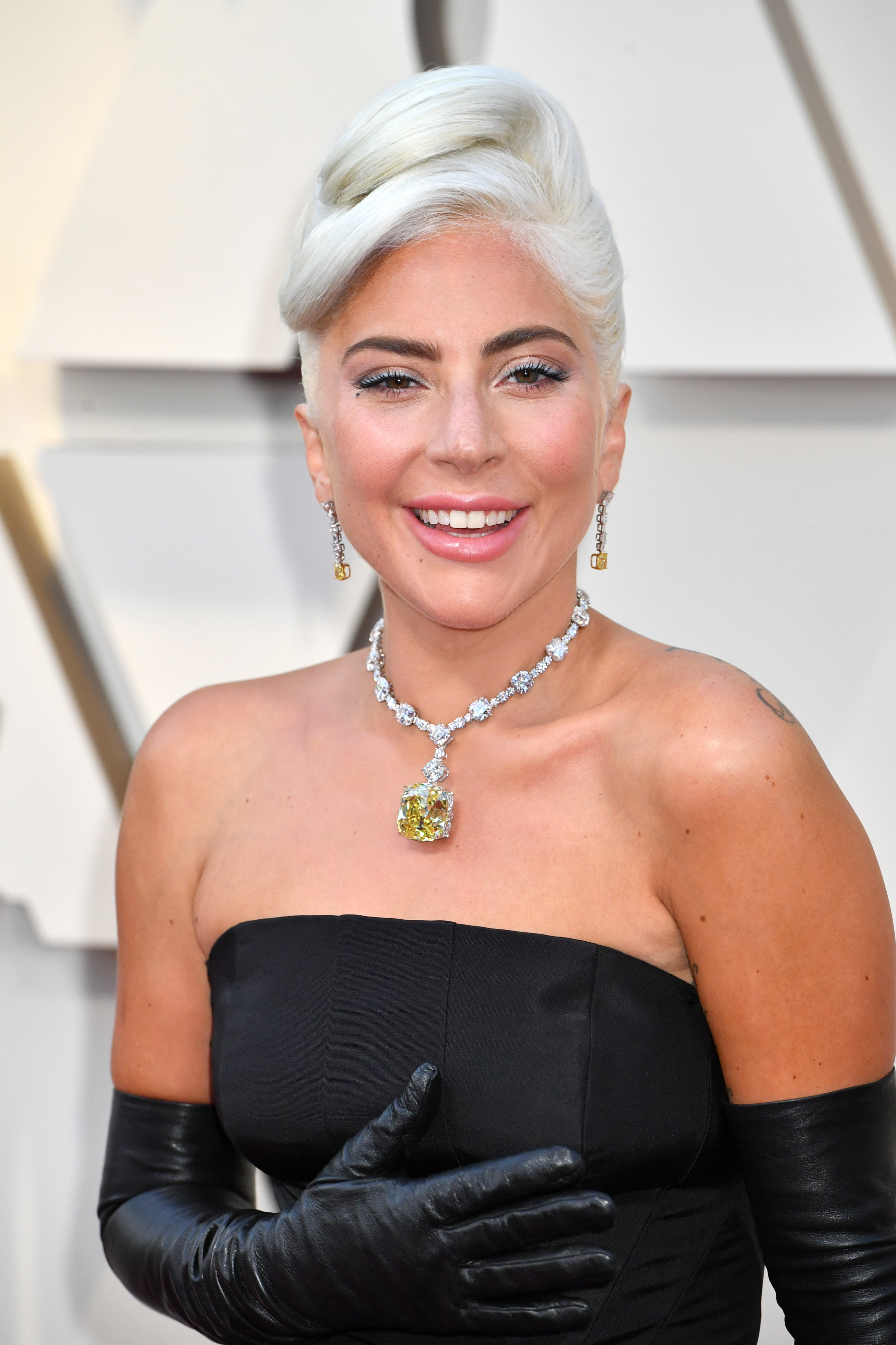 Lady Gaga smiling in a large jeweled necklace, gloves, and a gown