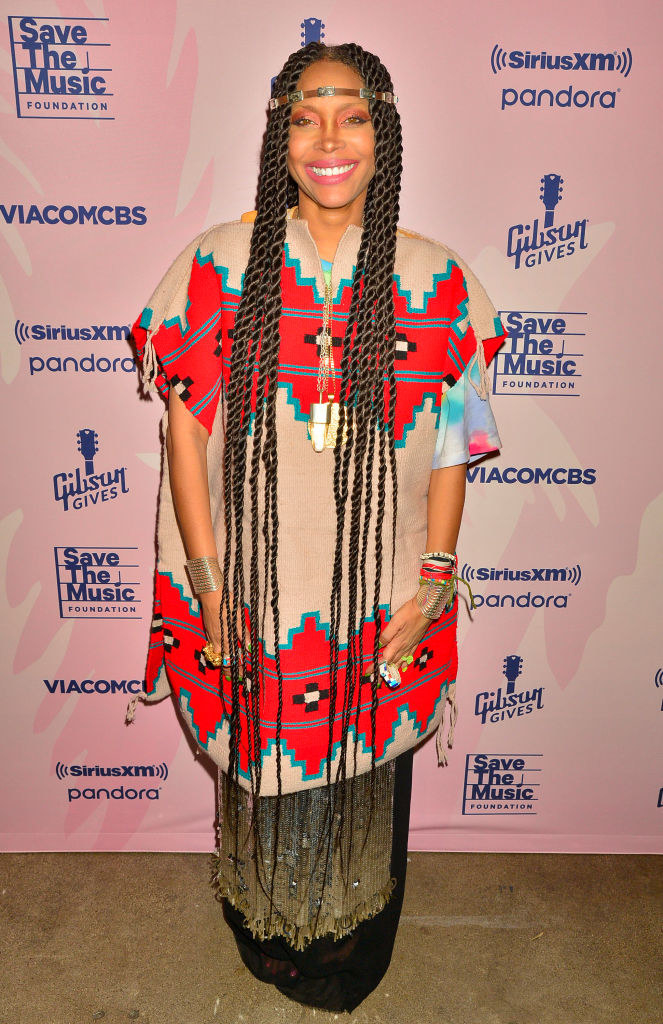 Erykah Badu stands in front of a step and repeat in almost floor-length braids and layered patterned dresses