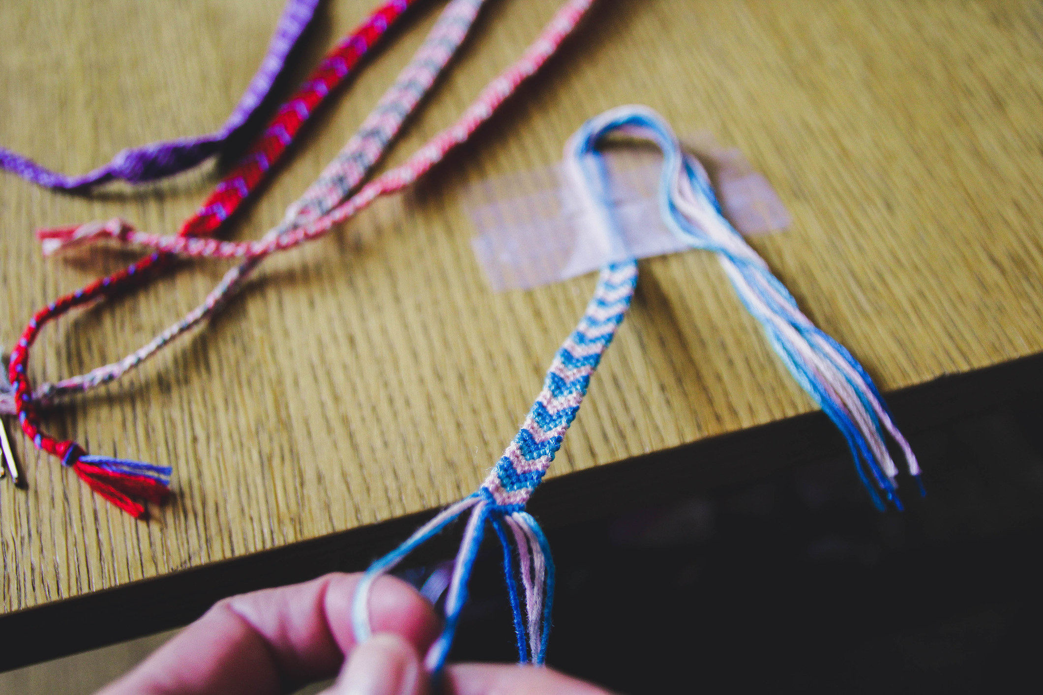 Hands making a friendship bracelet that&#x27;s taped to a desk and a bunch of bracelets next to it