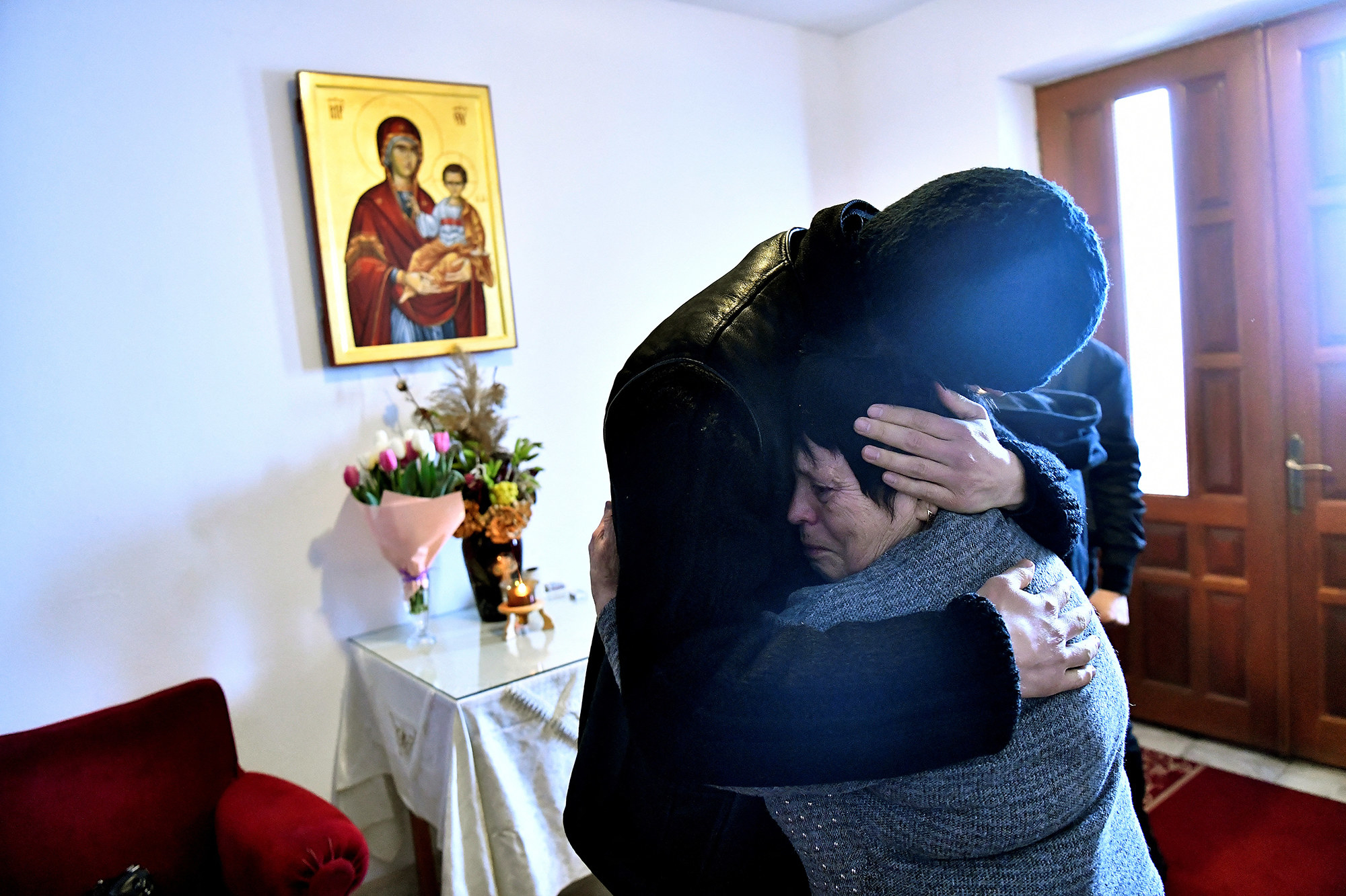 Father Gherasim Soca embraces Svetlana, a 75-year-old refugee, as she cries in a room with flowers on a table and a painting of an Orthodox saint above it