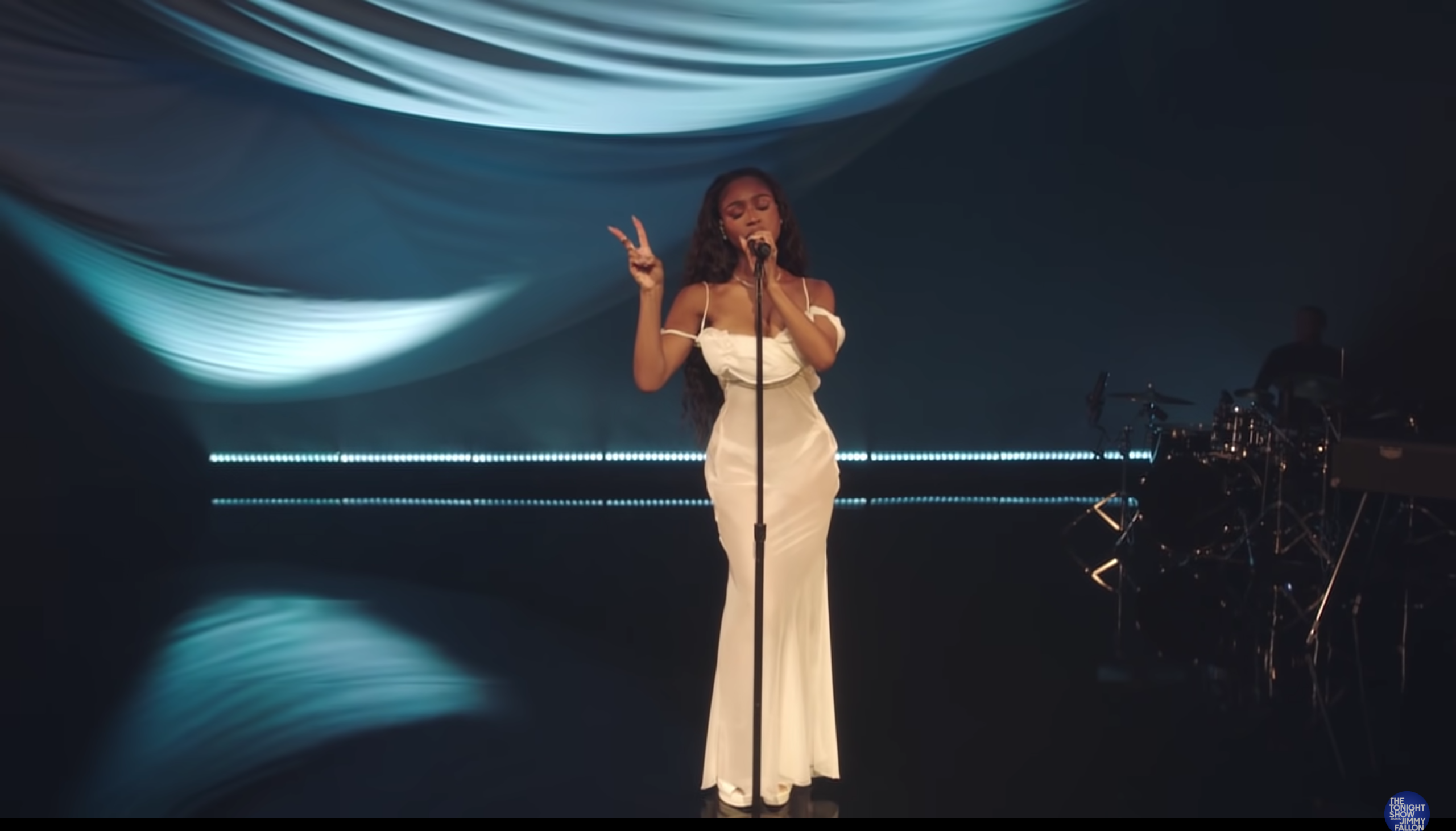 Normani in an all white dress singing her new song while doing a peace sign