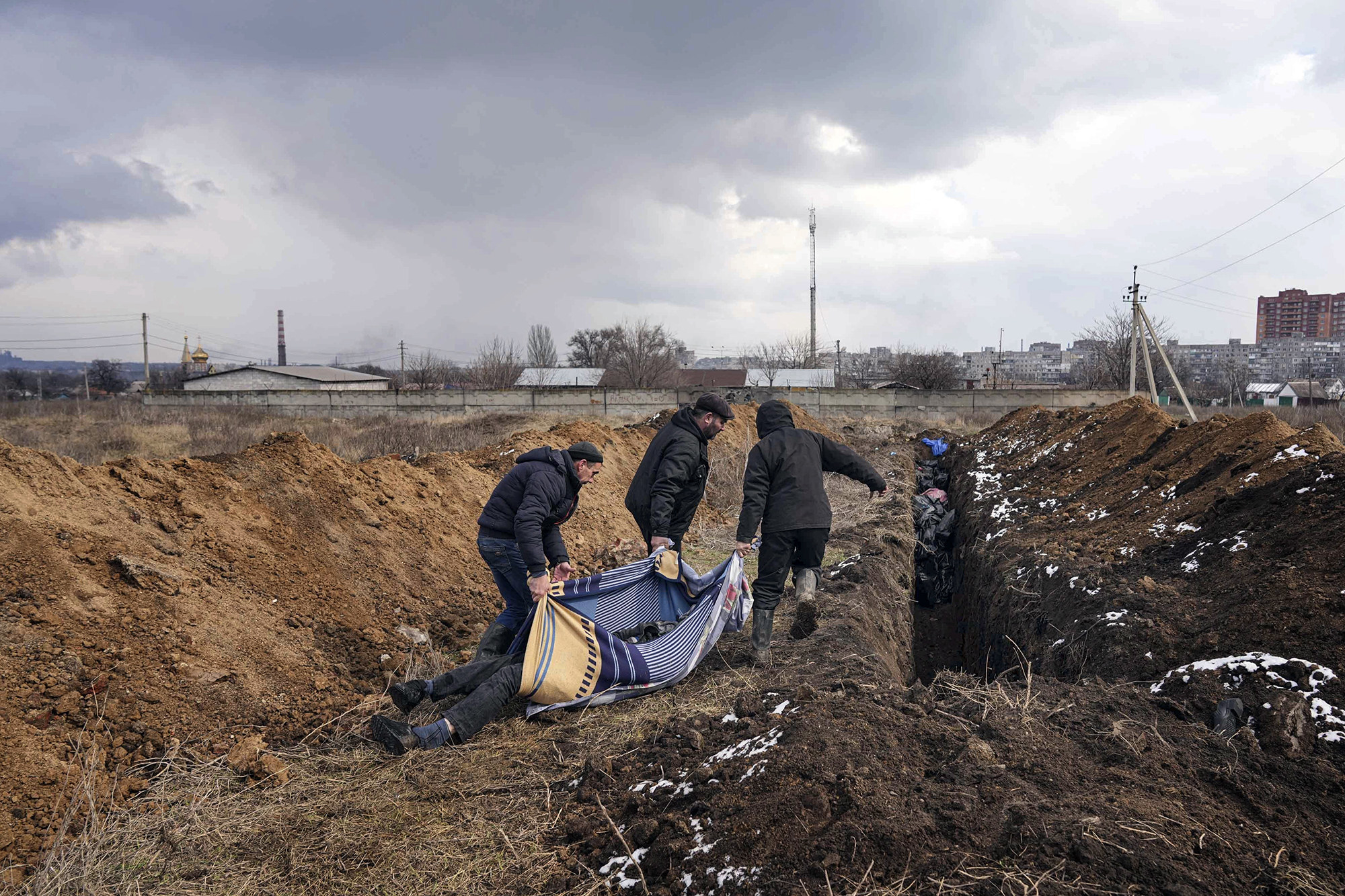 Three people carry a body in a blanket along a path toward a steep hole in the ground