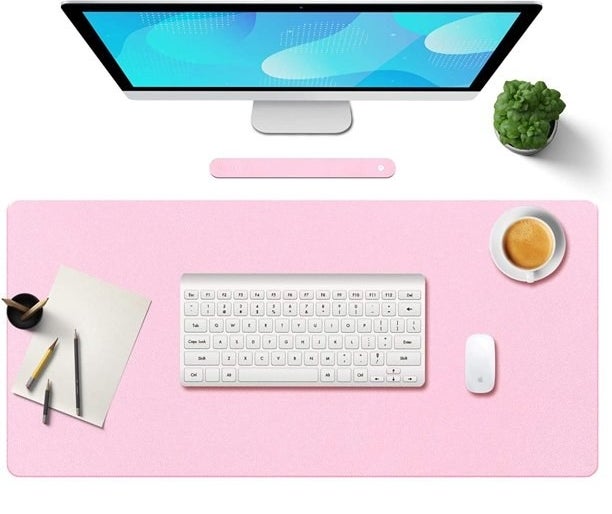 Pink desk in front of computer monitor with white keyboard, mouse, pen, paper, coffee cup on it