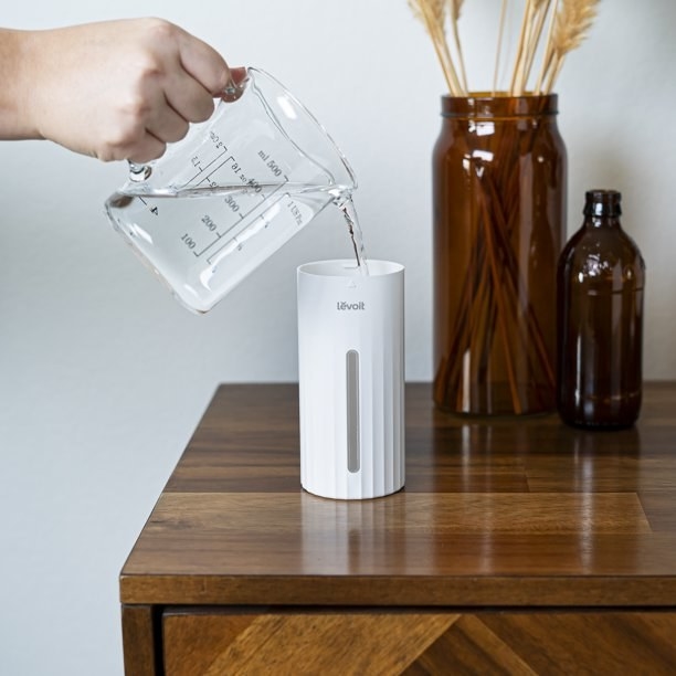 Model pouring water into white cylindrical humidifier on wooden table