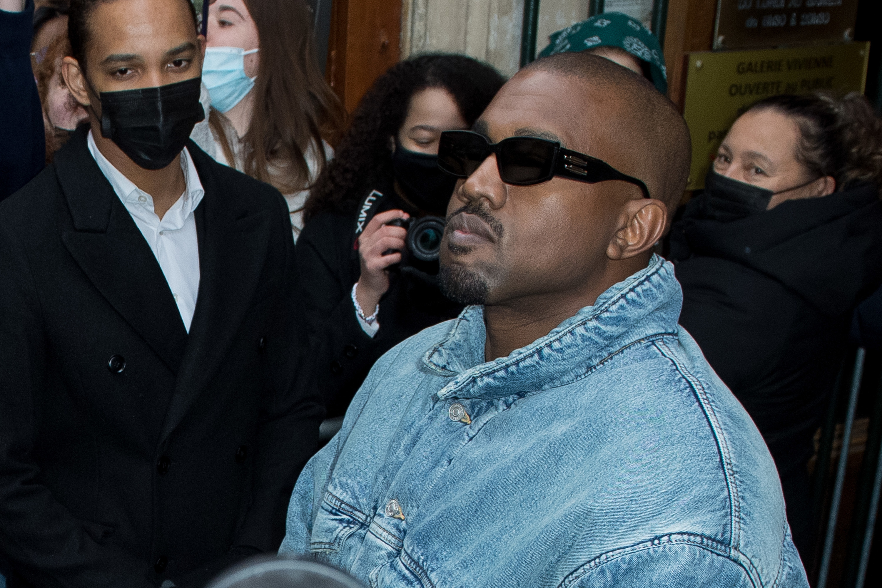 Ye in sunglasses and a jeans jacket with masked people behind him