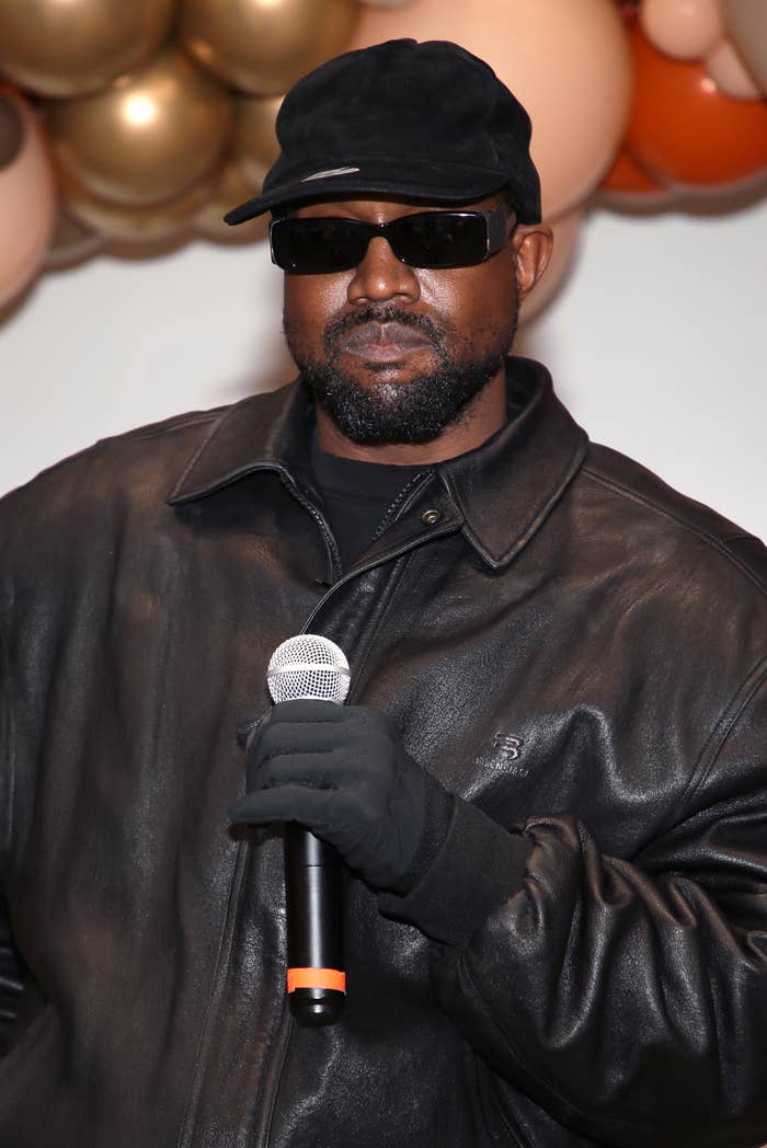 Ye, wearing a cap, sunglasses, and leather jacket, holds a microphone