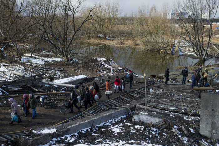 A number of people stand and walk by rubble near a body of water, with some snow on the ground