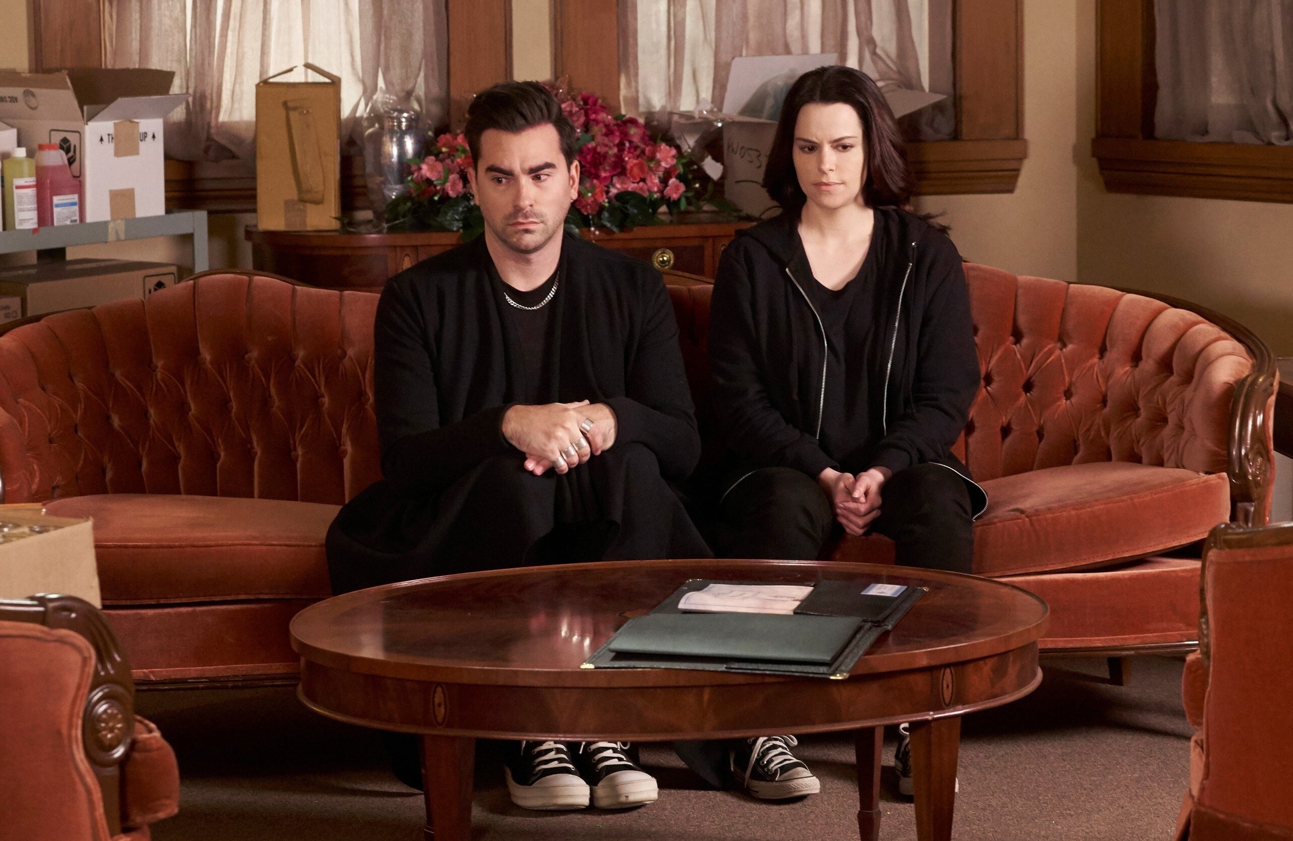 A man and a woman dressed in black sit on an orange couch.