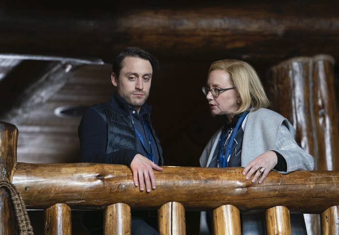 A man and an older woman stand in an indoors balcony looking down.