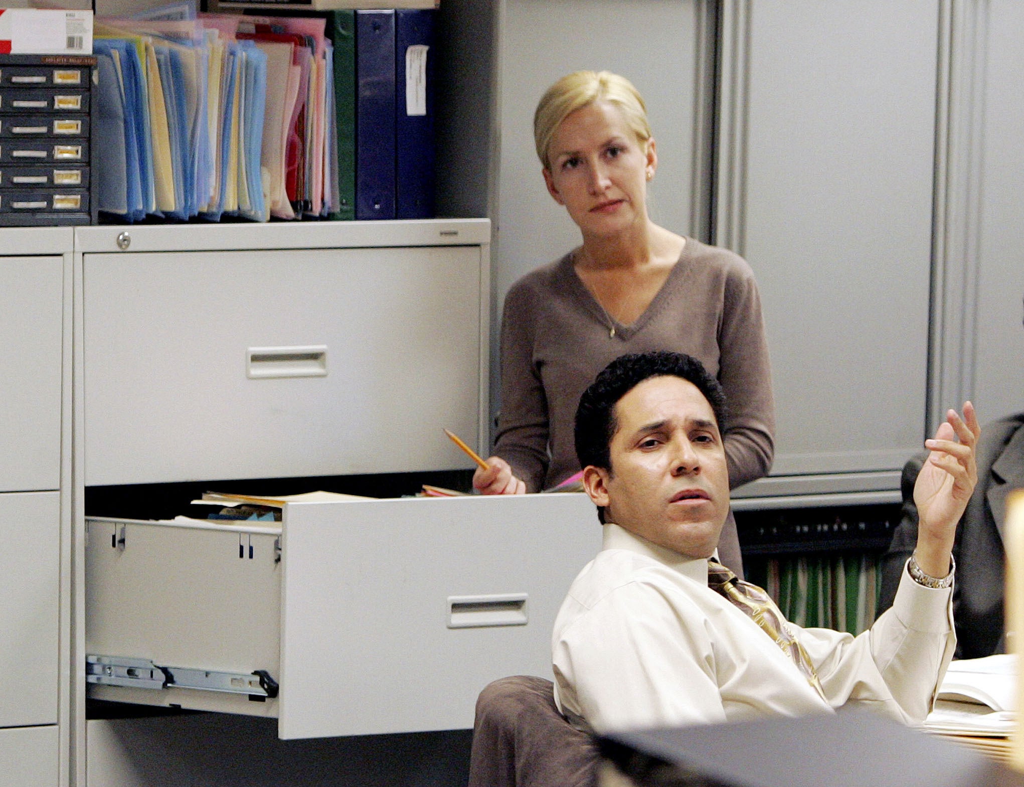 A woman stands at a filing cabinet. A man sits as a desk, he looks confused.