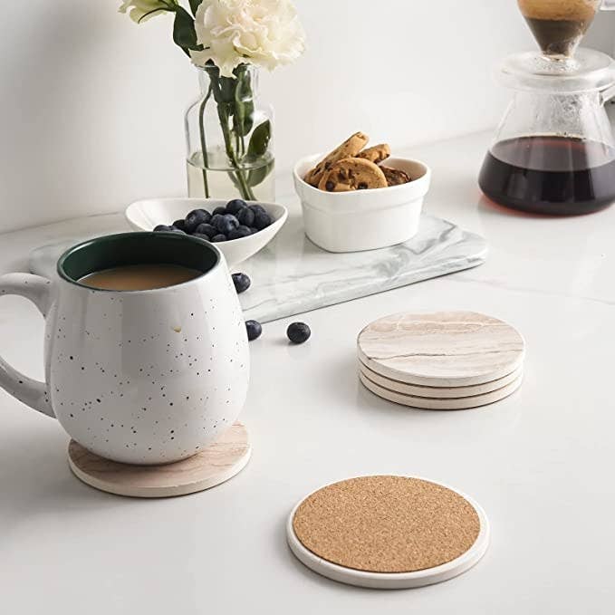 A mug full of tea sitting on a coaster in a kitchen with snacks behind it, and a stack of coasters beside it