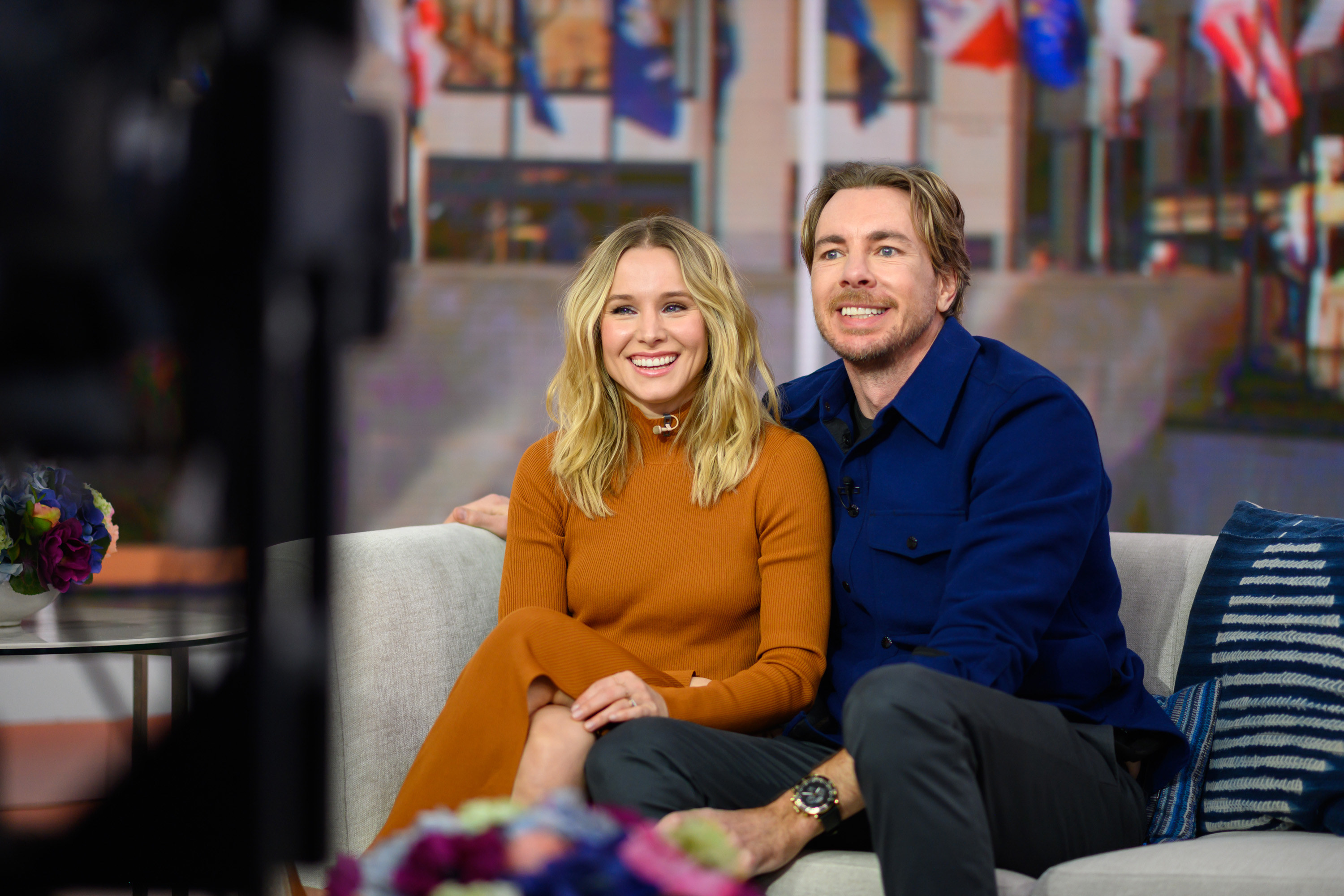 Dax with his arm around Kristen as they sit on a couch with cameras pointing to them