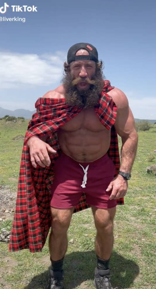A bearded man standing in a field wearing boots, swim trunks, a plaid cape records a TikTok video