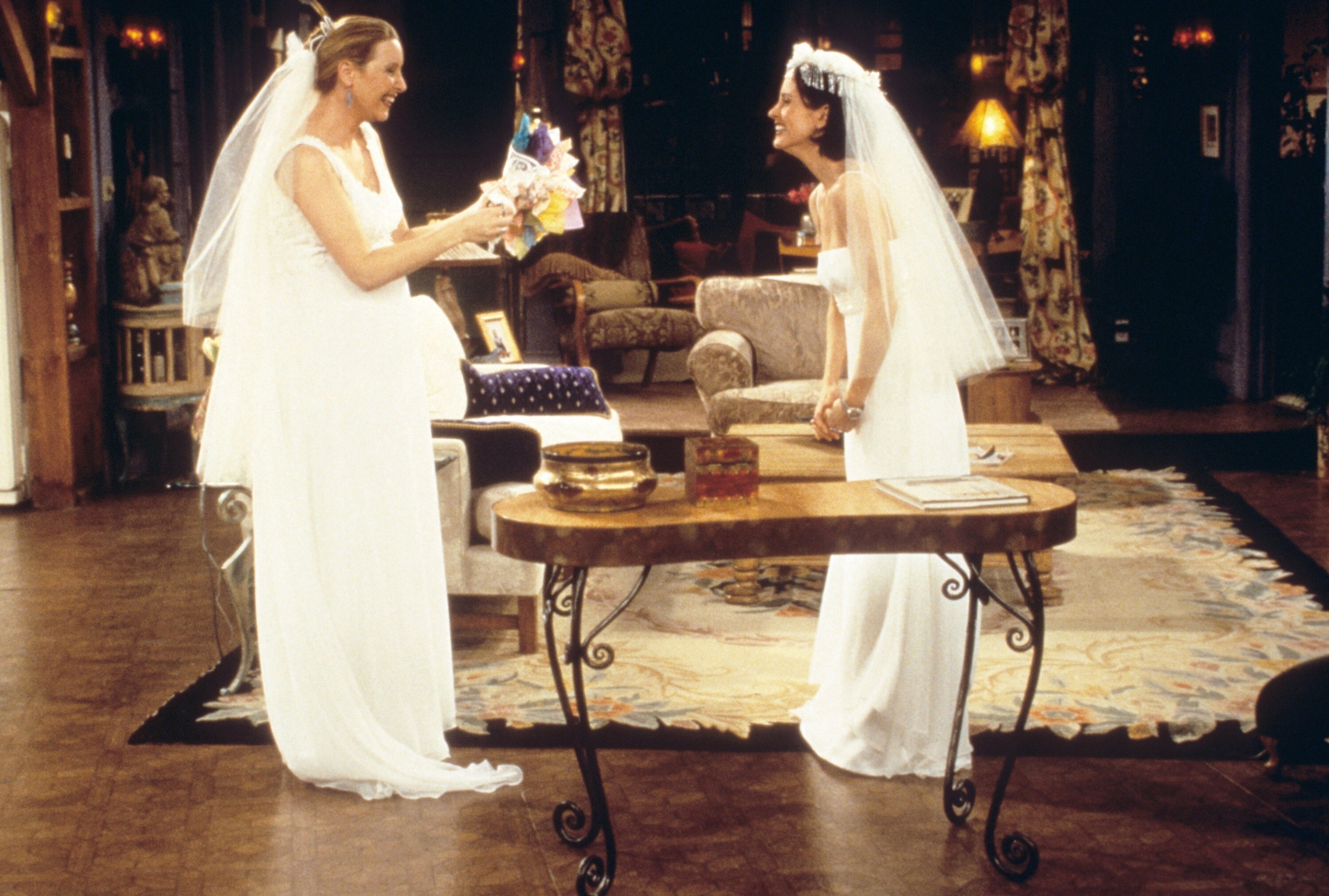 Two women stand in a living room, both in wedding dresses.