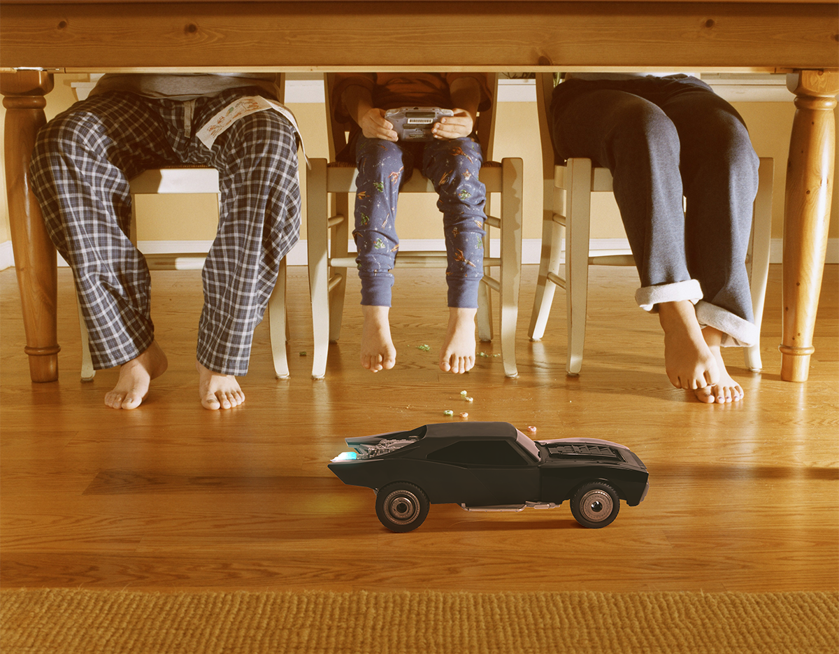 Under the dining room table, we see the Turbo Boost Batmobile™ RC and a father&#x27;s and kids&#x27; legs.