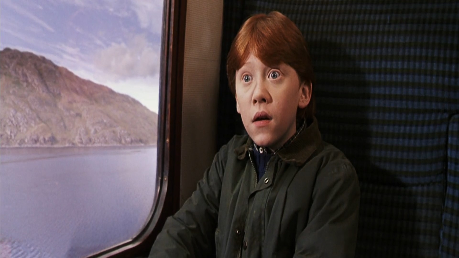 Rupert Grint is sitting in a train compartment and looks in surprise at someone