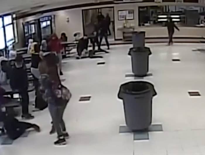 Footage of school lunchroom showing a number of students standing around and, in the background, someone lying on the ground with an adult kneeling over them