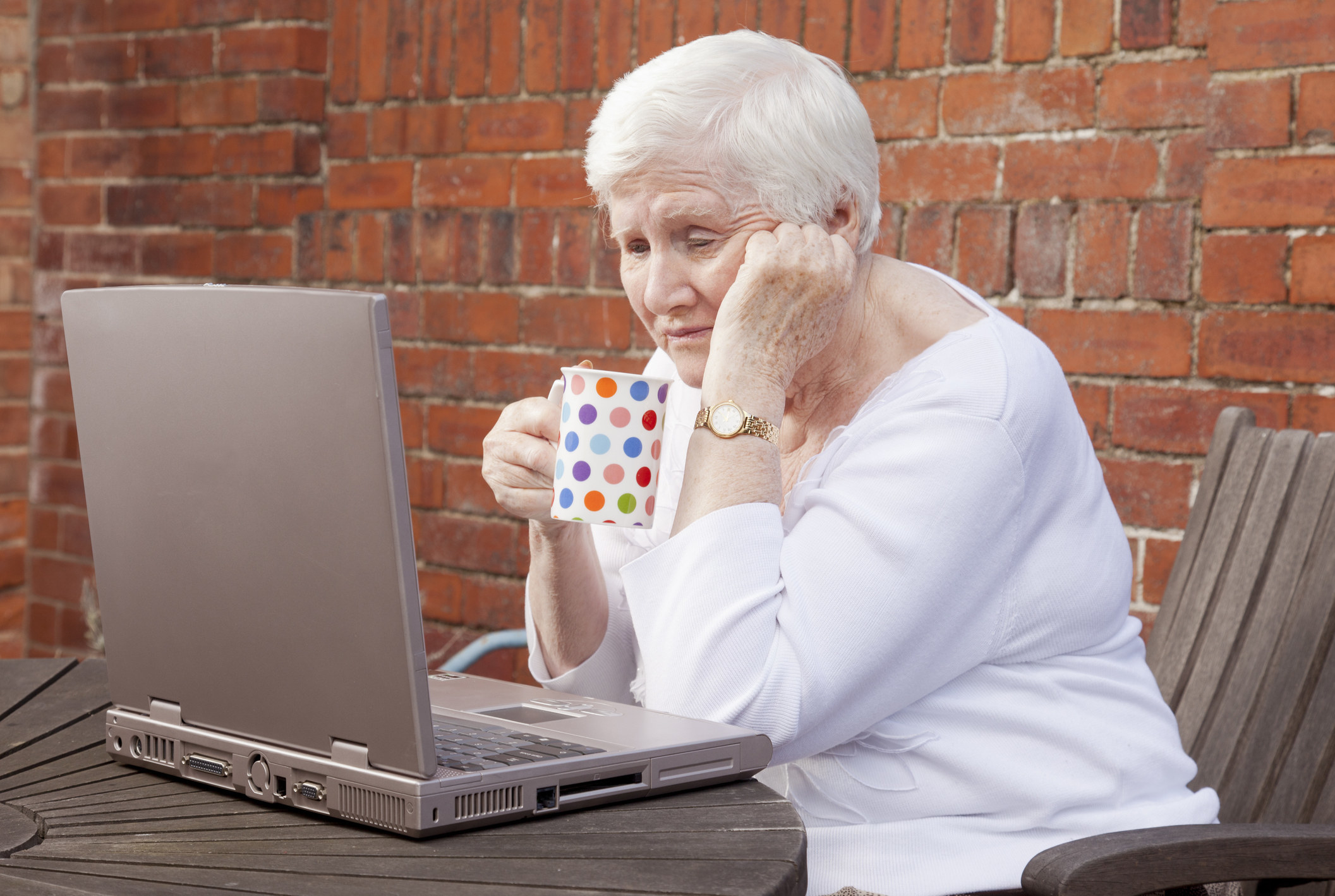 Older woman sitting at a laptop holding a mug and looking absorbed