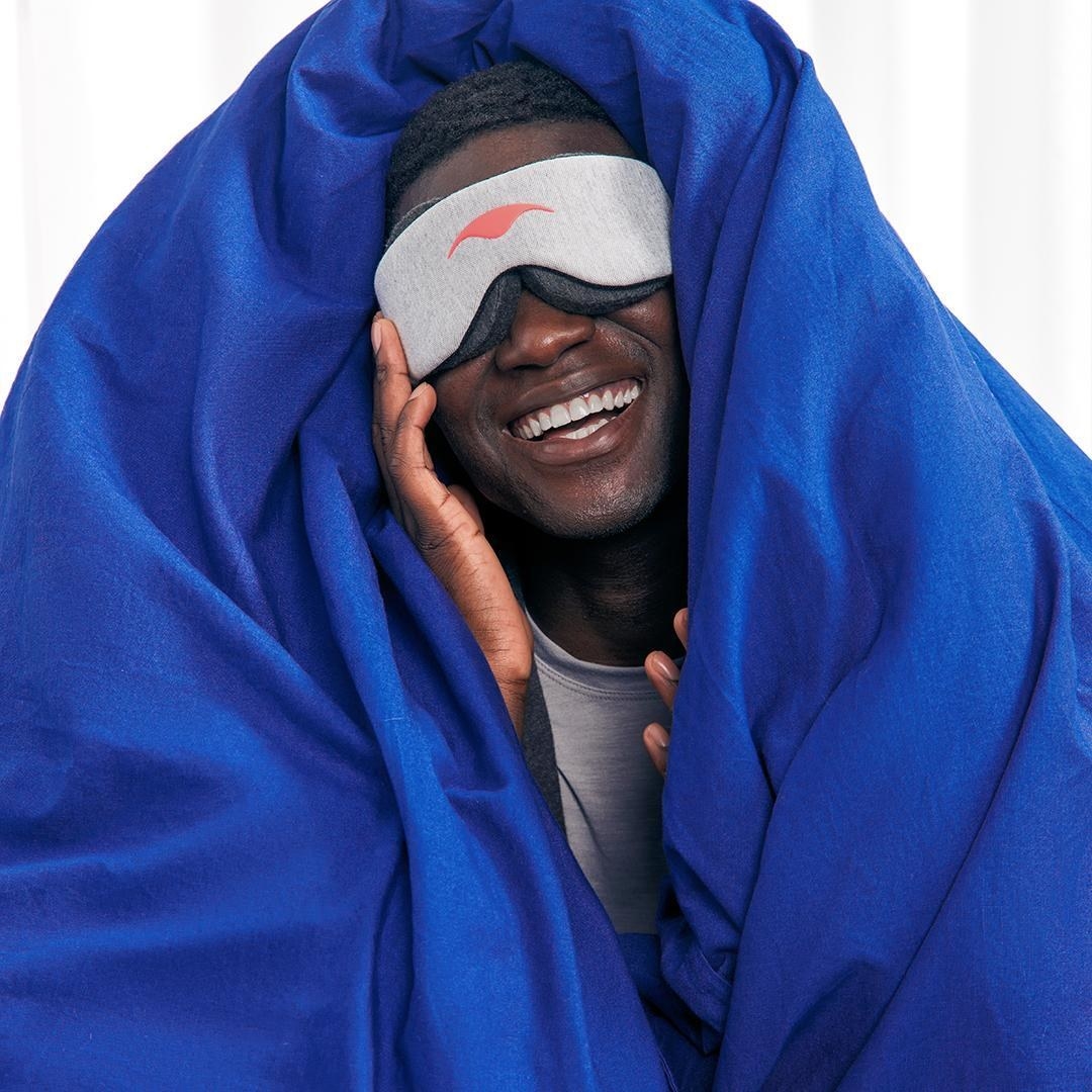 a smiling person wrapped in a blanket while wearing the contoured eye mask