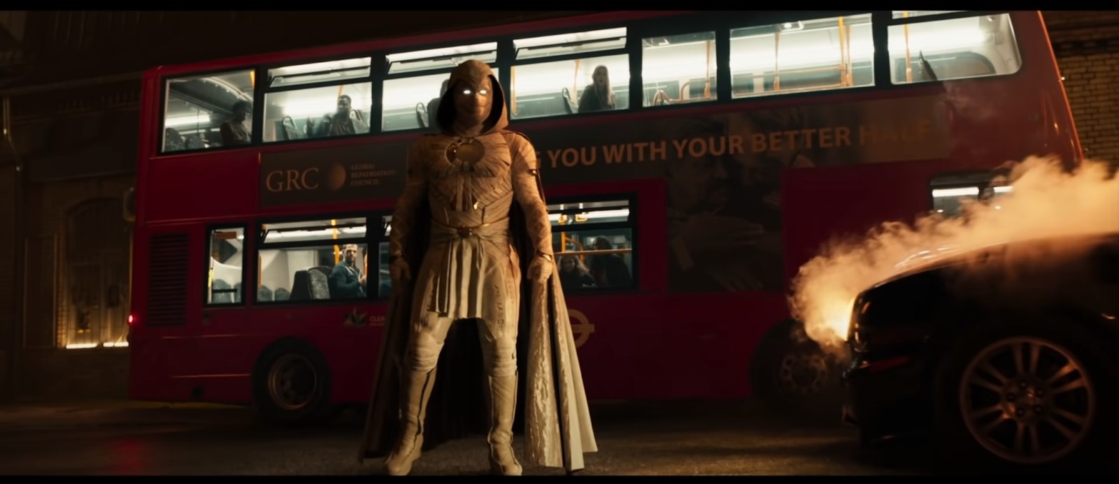 Moon Knight standing in front of a red double decker bus with a sign for the GRC on it
