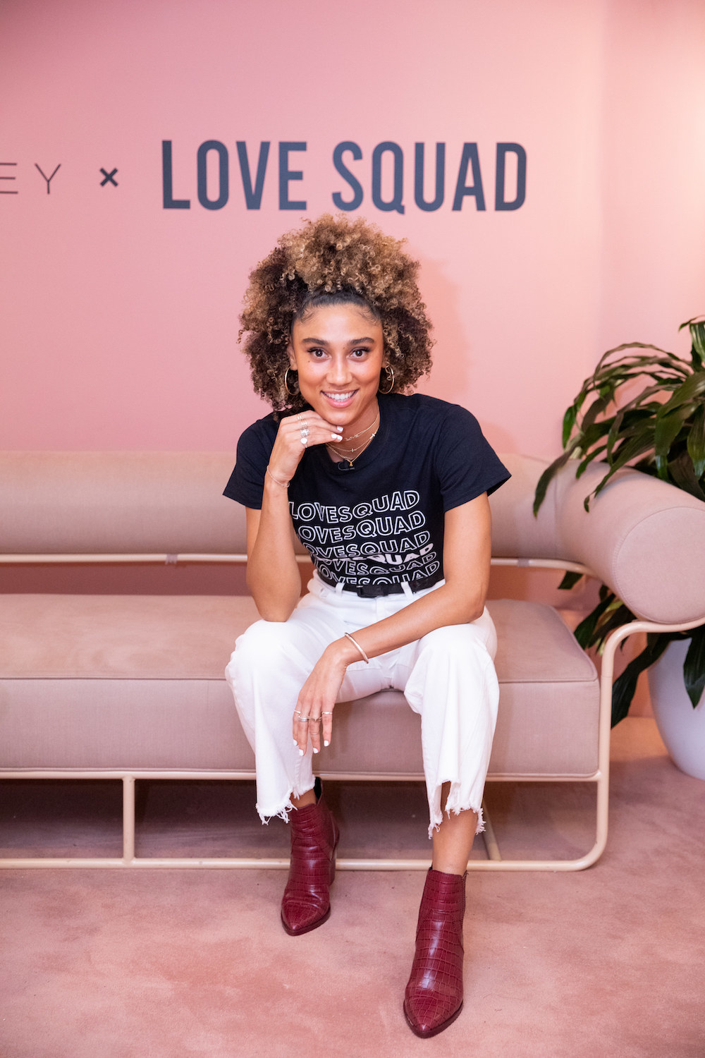 A woman wearing a shirt that says &quot;Love Squad&quot; sits in front of a wall that reads &quot;Love Squad&quot; and smiles for the camera