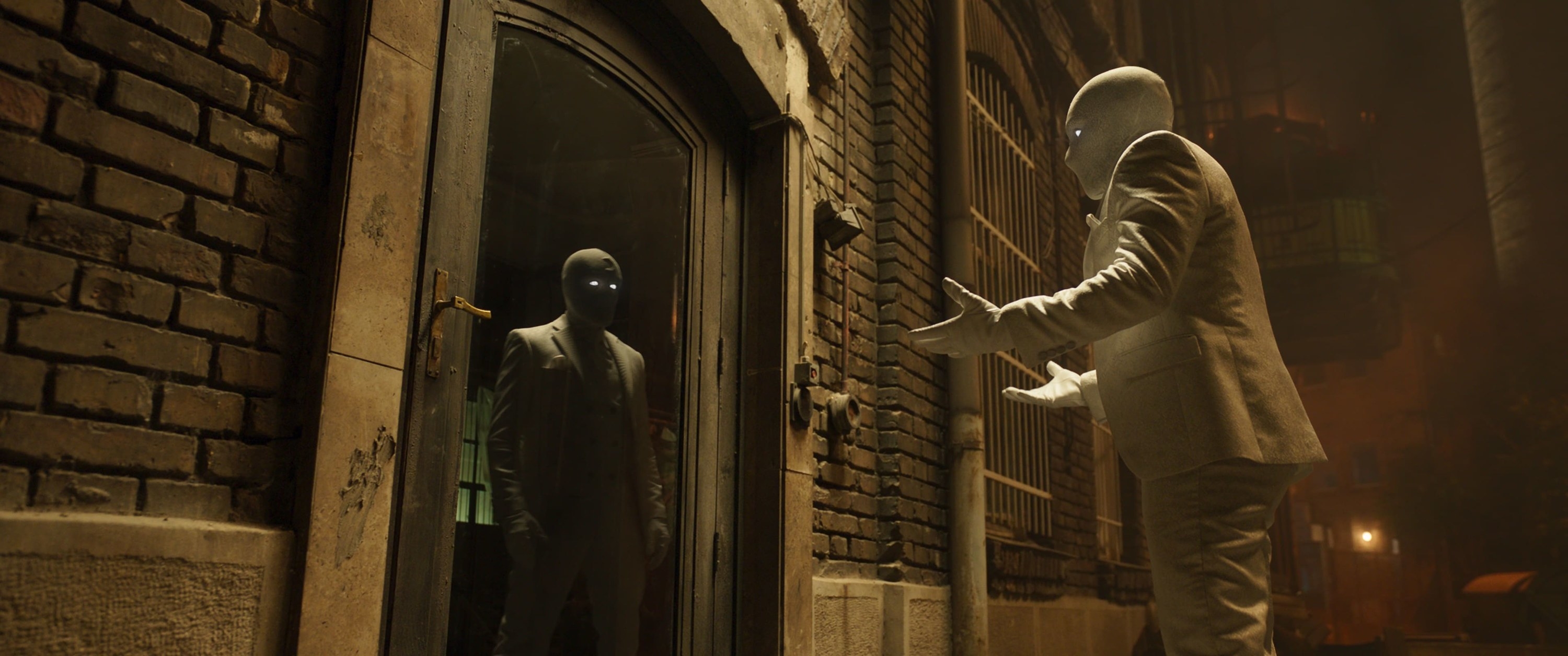 Mr Knight, dressed in a white suit with his white headgear on, gestures at himself in the reflection of a glass door. He is standing on a street, and his reflection is NOT mirroring his actions