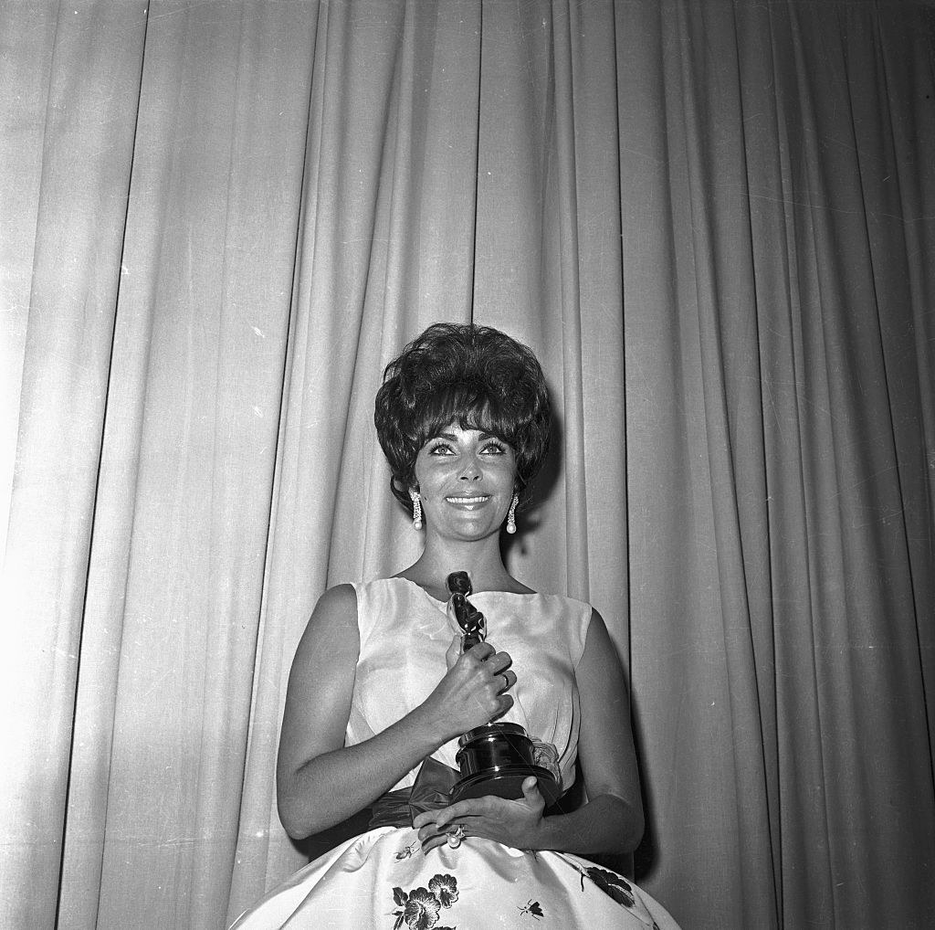 Elizabeth wears a sleeveless dress and short hair as she poses with her award