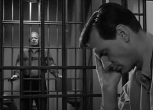Foreground: A man is seated behind jail bars with his face in his hand. Background: A man is standing behind jail bars and facing the man in the foreground