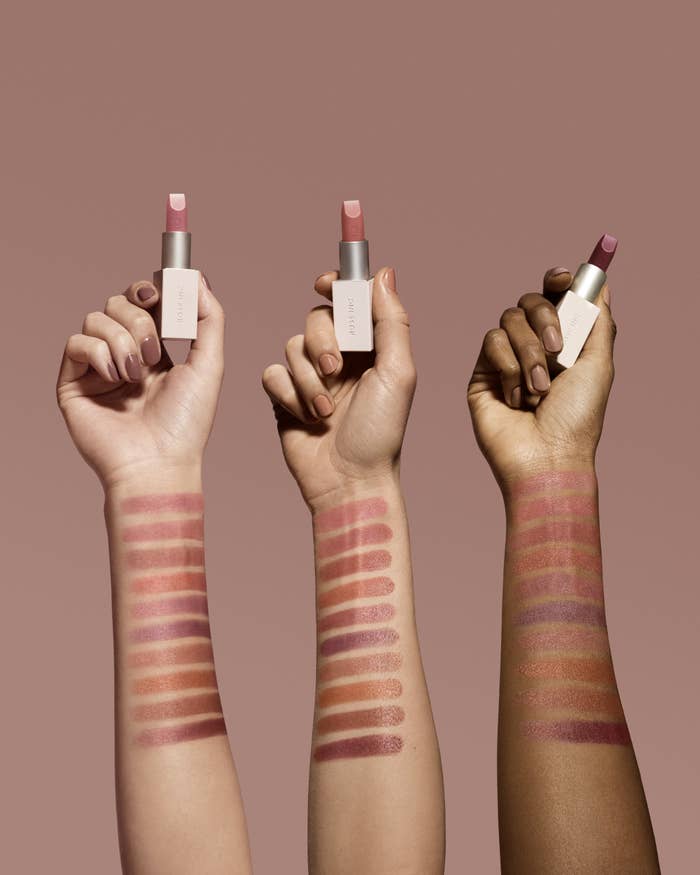 Three arms of different skin tones hold lipsticks, and the arms each are smeared with several lines of different colors of lipstick