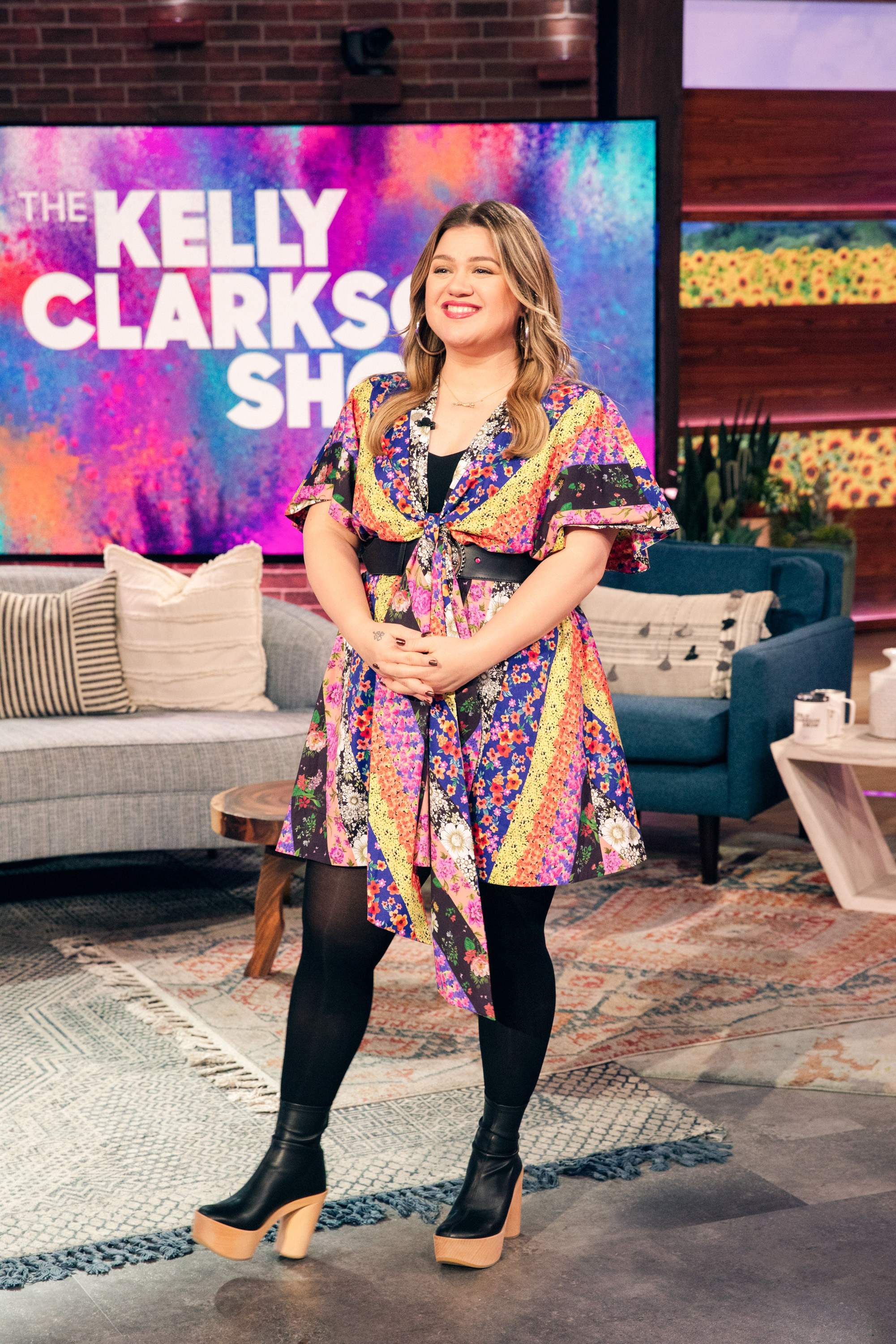Kelly smiles while standing on the set of her daytime talkshow The Kelly Clarkson Show