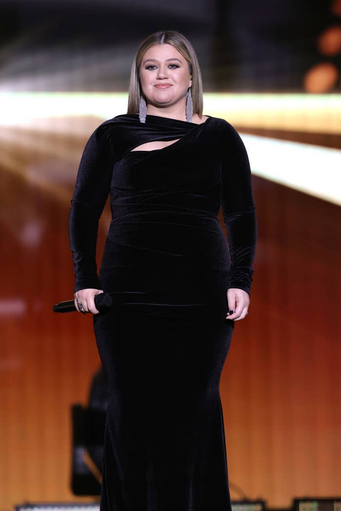 Kelly smiles while holding a microphone and looking out at the audience while wearing a velvet column gown