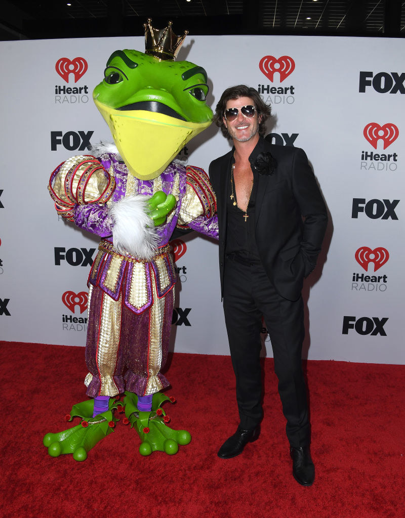 Robin wearing his shirt partially open, a blazer, and slacks as he poses on the red carpet with a person in a costume from the Masked Singer