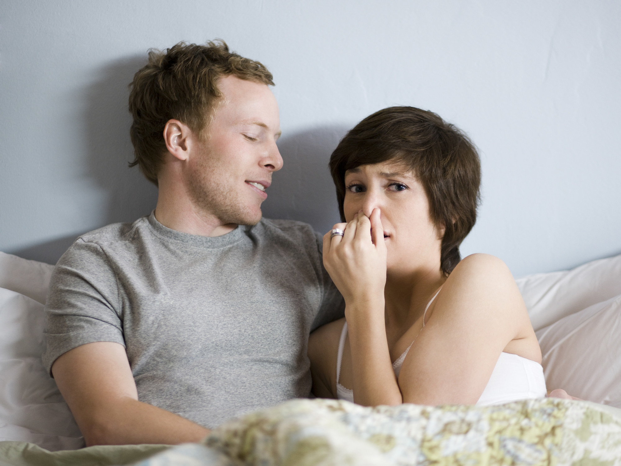 Una pareja en la cama y la mujer hace una señal con el rostro de que siente un mal olor.