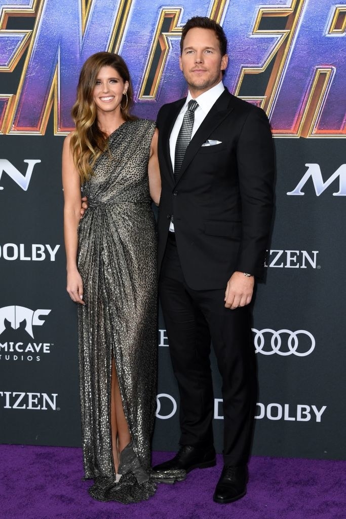 on the red carpet, Katherine wears a floor-length one-shoulder dress with a sparkly pattern, and Chris wears a classic suit with a tie that matches her dress