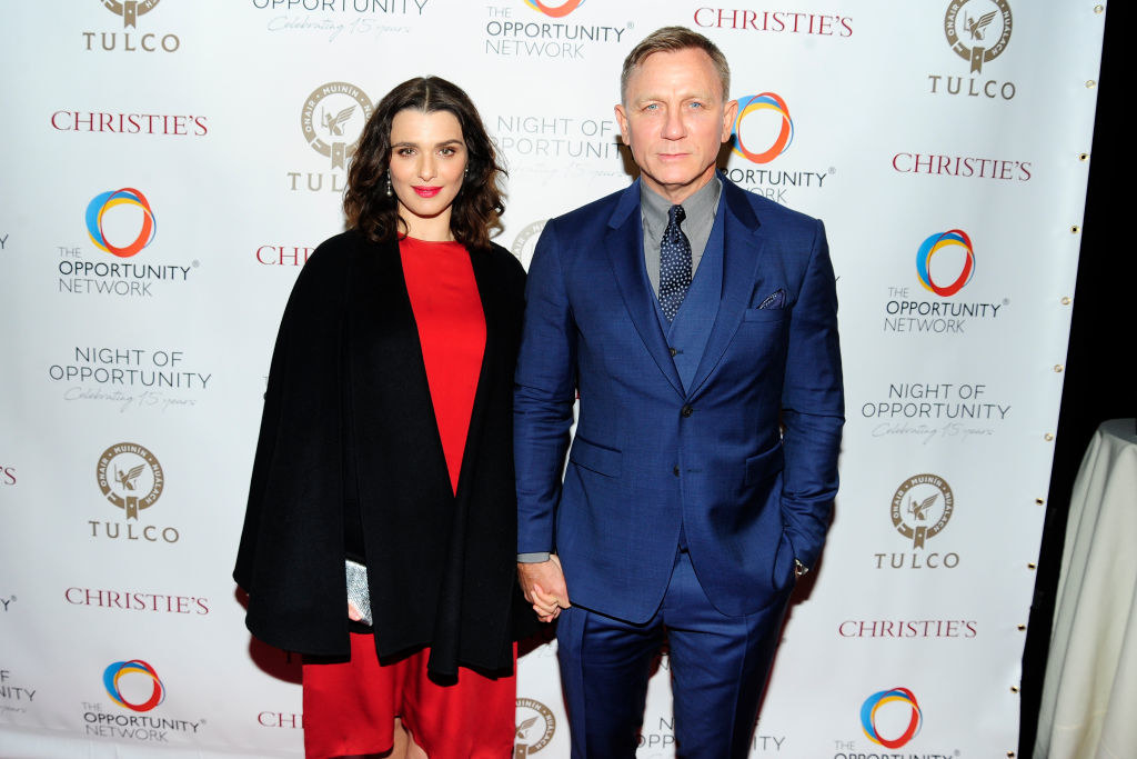 holding hands on the red carpet, Rachel wears an oversized cardigan over a bold pantsuit, and Daniel wears a simple suit in a complimentary bold color