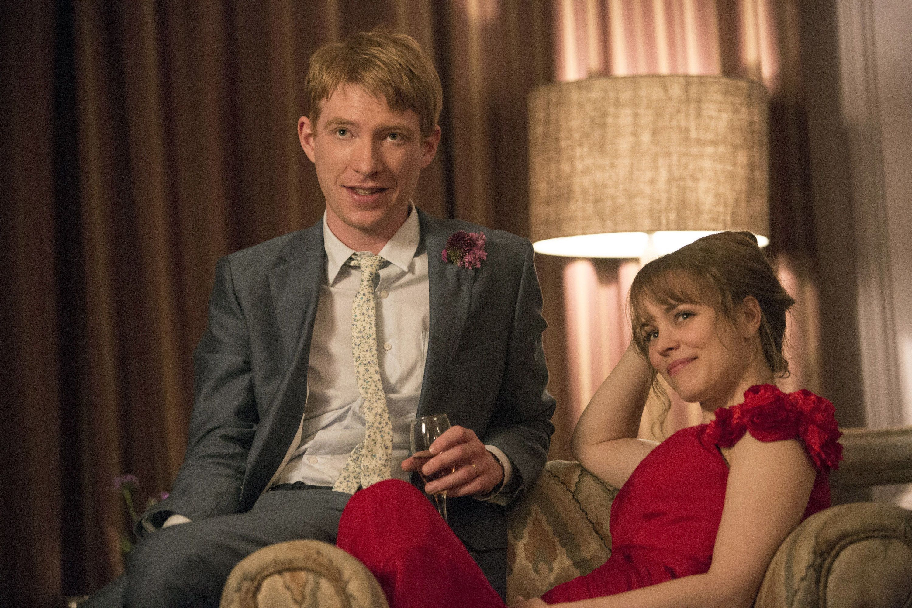 Domnhall Gleeson and Rachel McAdams, wearing a suit and dress, respectively, sit next to each other in a living room in a scene from About Time