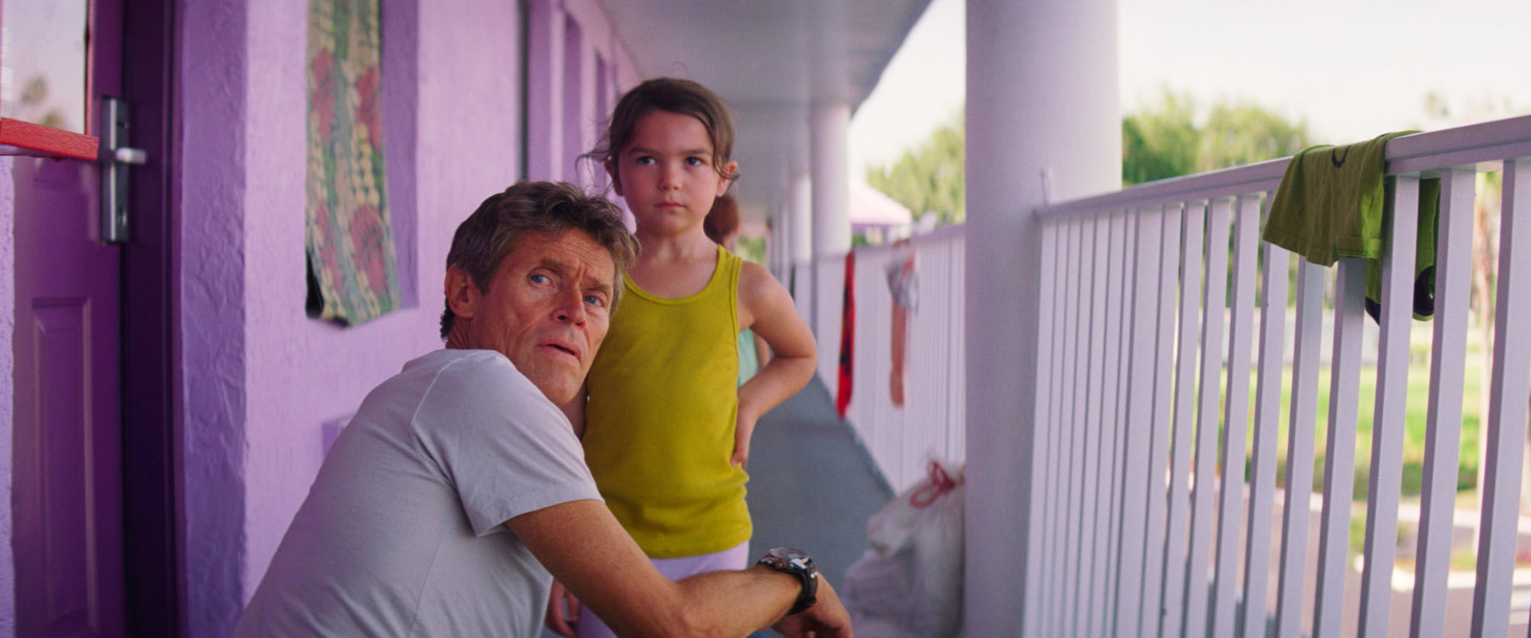 Willem Dafoe next to a little girl as he kneels on a balcony hallway in a scene from The Florida Project