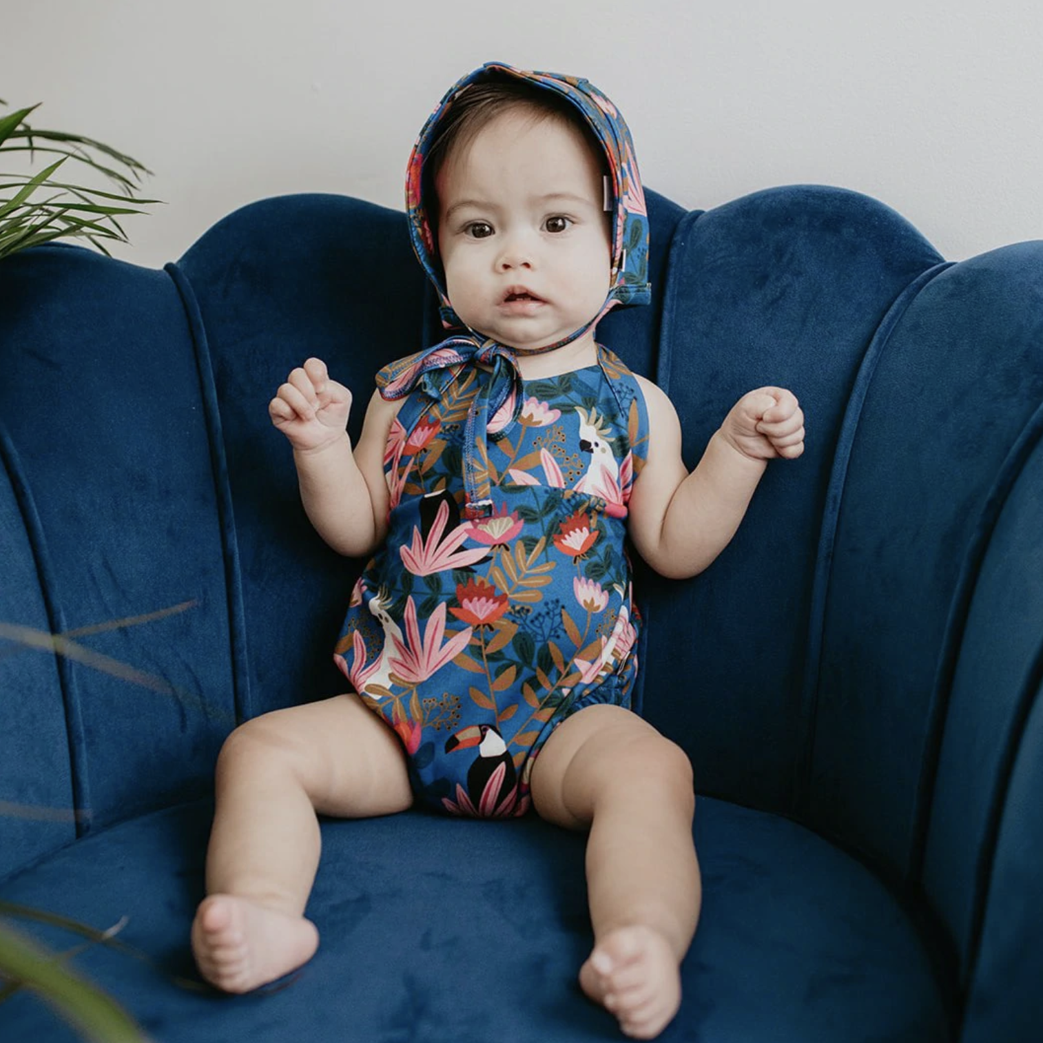 A baby sitting on a velour chair wearing the floral onesie and matching bonnet