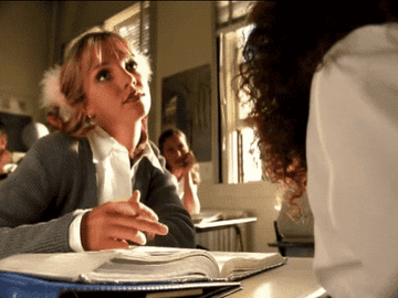 Britney Spears dressed as a school girl, hitting her pencil against a school book while she leans on one hand. She is sat at her desk in a classroom.