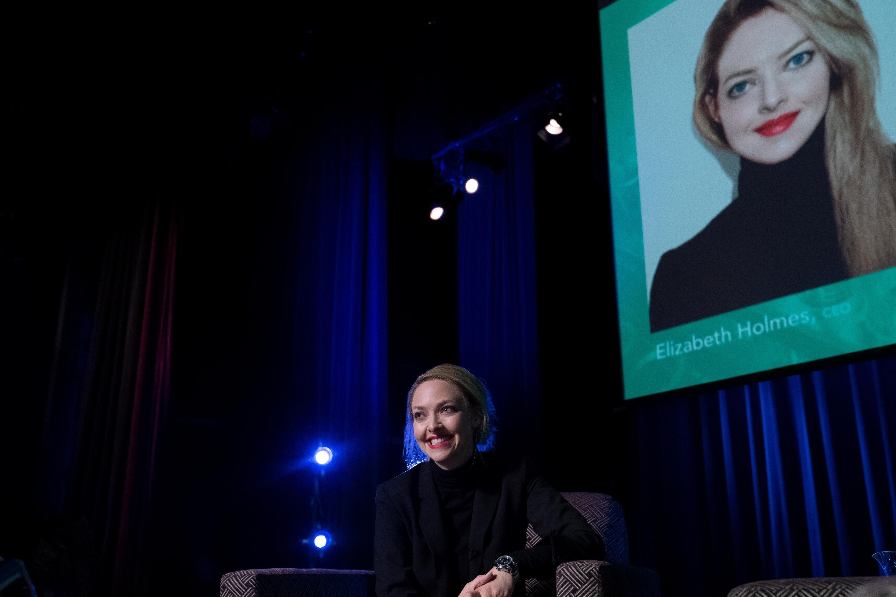 Seyfried smiles while sitting in a couch in front of a picture of Elizabeth Holmes